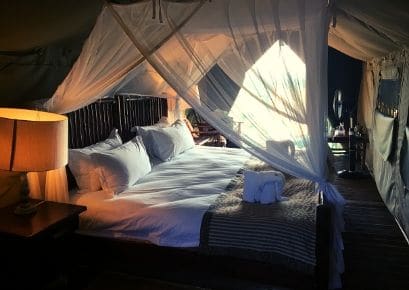 The interior of an Alexandra's Africa luxury safari tent - showing a cosy king size double bed, with mosquito nets and lamp on bedside table in the foreground