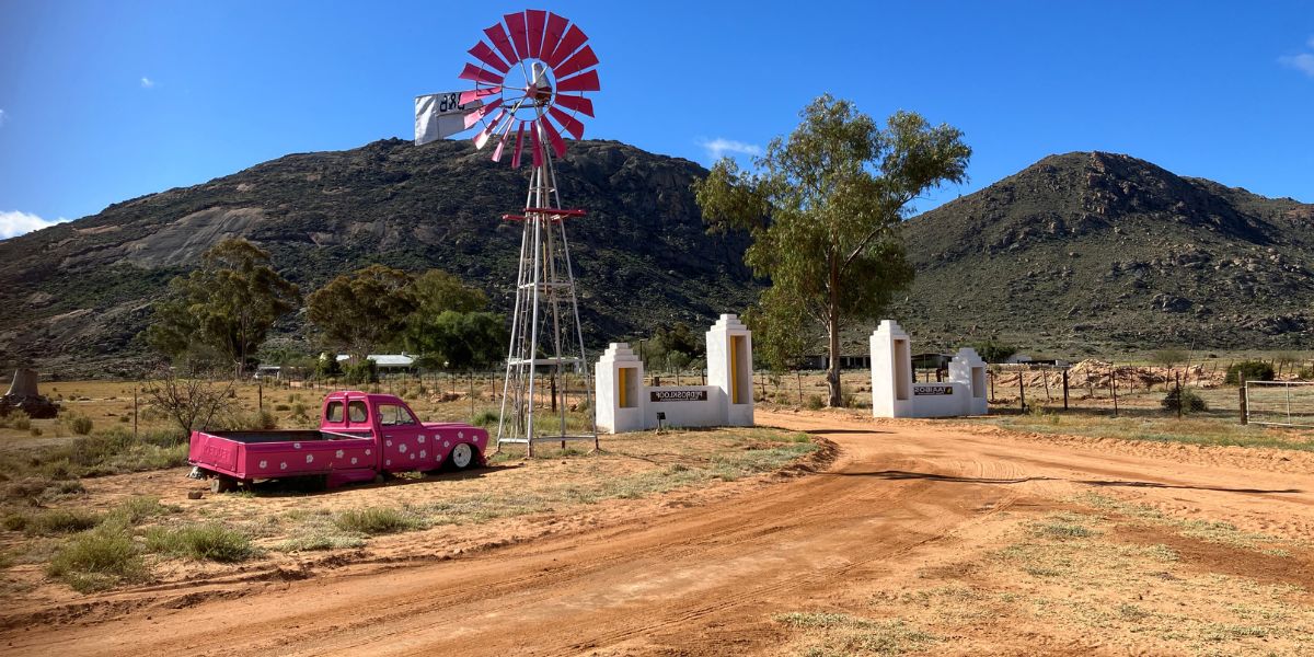 Picture of farm from outside white gates with pink car and wind pump in foreground