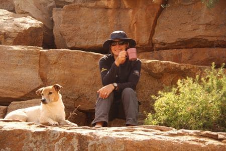 Woman sitting in rocky canyon drinking coffee with alert dog lying on rocks in foreground