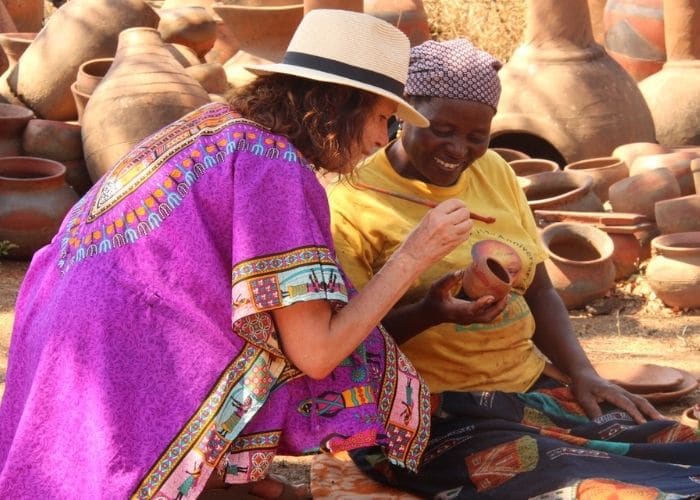 Alexandra's Africa guest kneeling next to Venda lady, all in bright clothes, being taught how to paint a Venda clay pot
