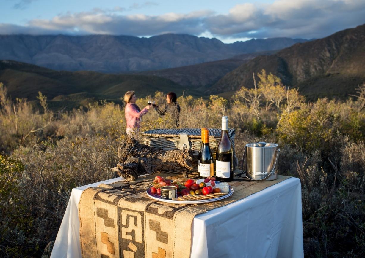 Small table covered by tablecloth with drinks and snacks in foreground and in background two women with glasses with mountains and bushveld in the distance at sundown
